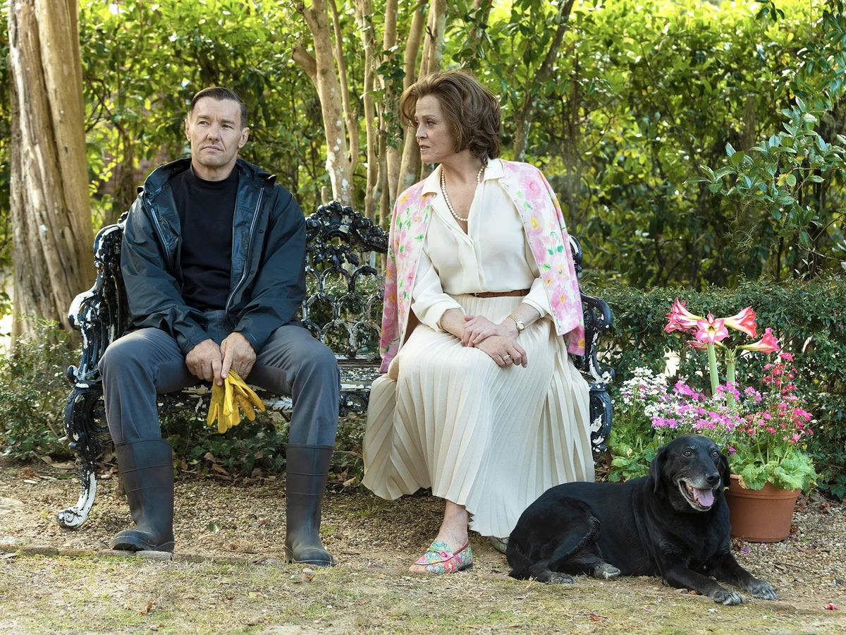 (L-R) Joel Edgerton and Sigourney Weaver sitting on a bench near an arrangement of potted flowers and a black dog in Master Gardener.