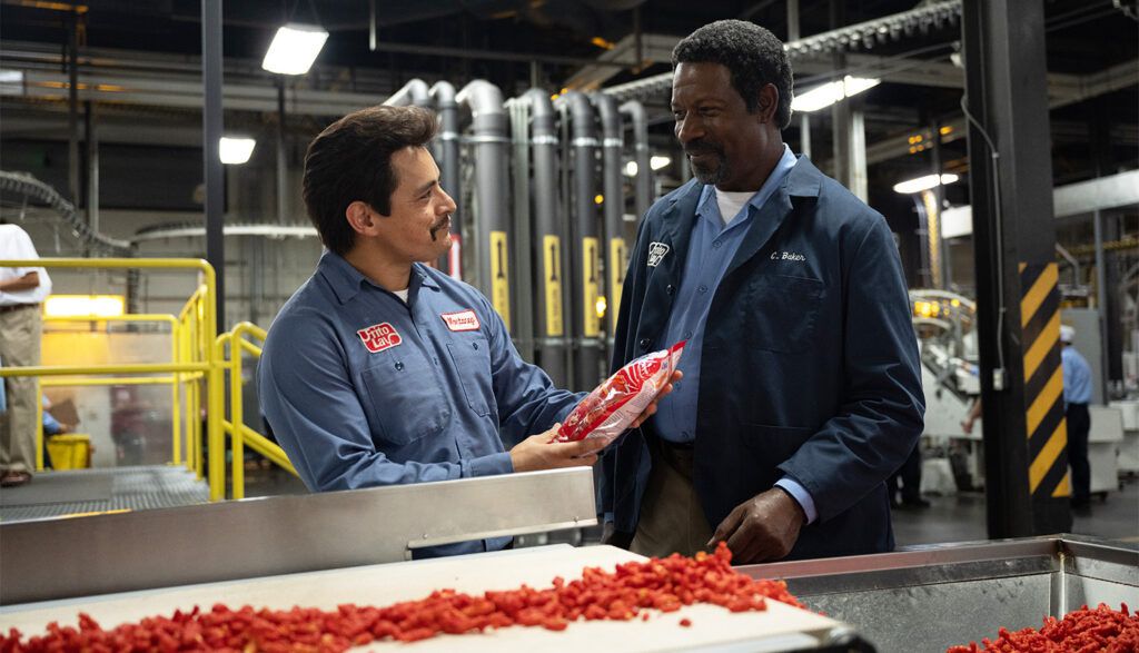 (L-R) Jesse Garcia and Dennis Haysbert holding a bag of Flamin’ Hot Cheetos in front of a factory line of Cheetos in Flamin’ Hot.