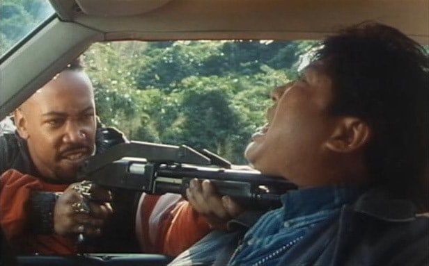 Robert Samuels points a shotgun at Sammo Hung’s neck from outside a car in Don’t Give a Damn.