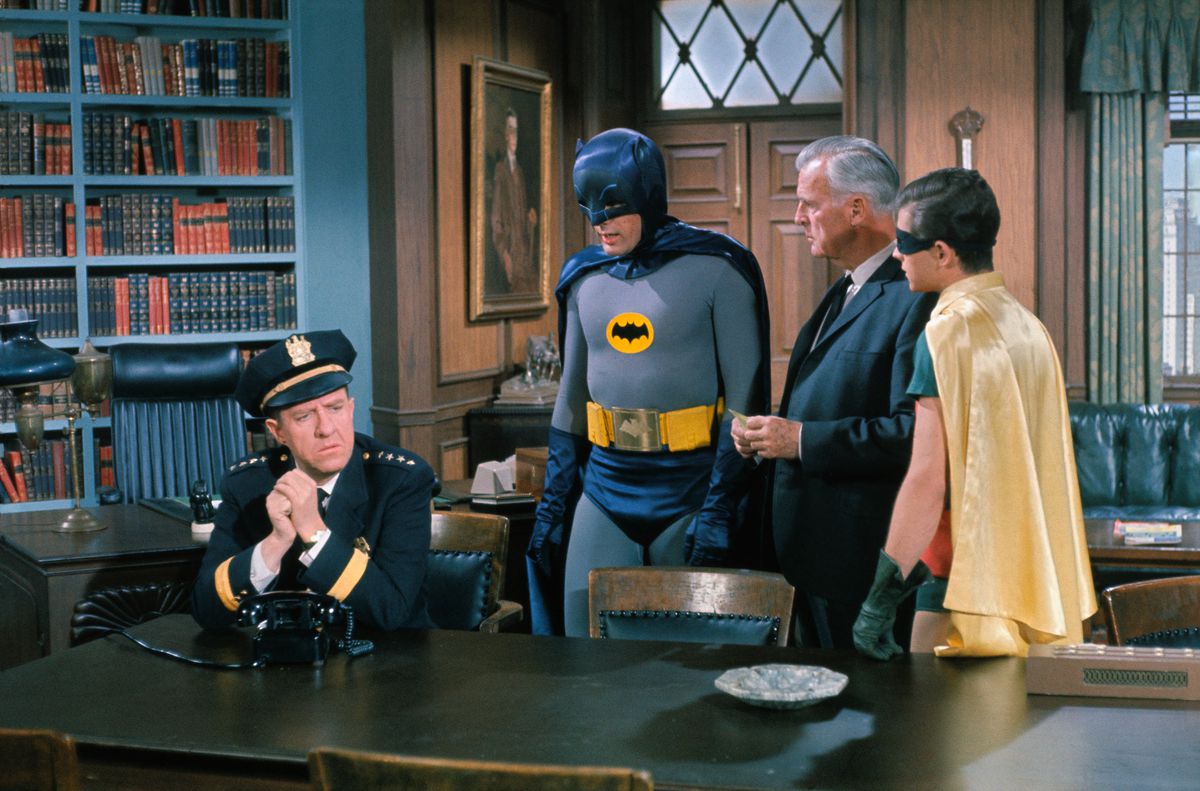 Adam West och Burt Ward som Batman och Robin, står i ett bibliotek och pratar med kommissarie Gordon och chefen O'Hara i Batman (1966).