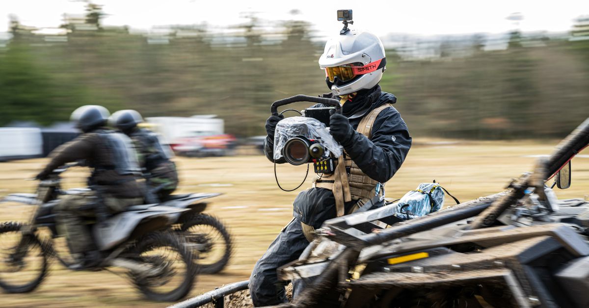 Regissören Sam Hargrave håller i en motorcykelhjälm, skyddsglasögon och en säkerhetssele.  upp en kamera och bär en andra på huvudet, sitter på framsidan av ett fordon i rörelse medan han filmar en sekvens i Extraction 2