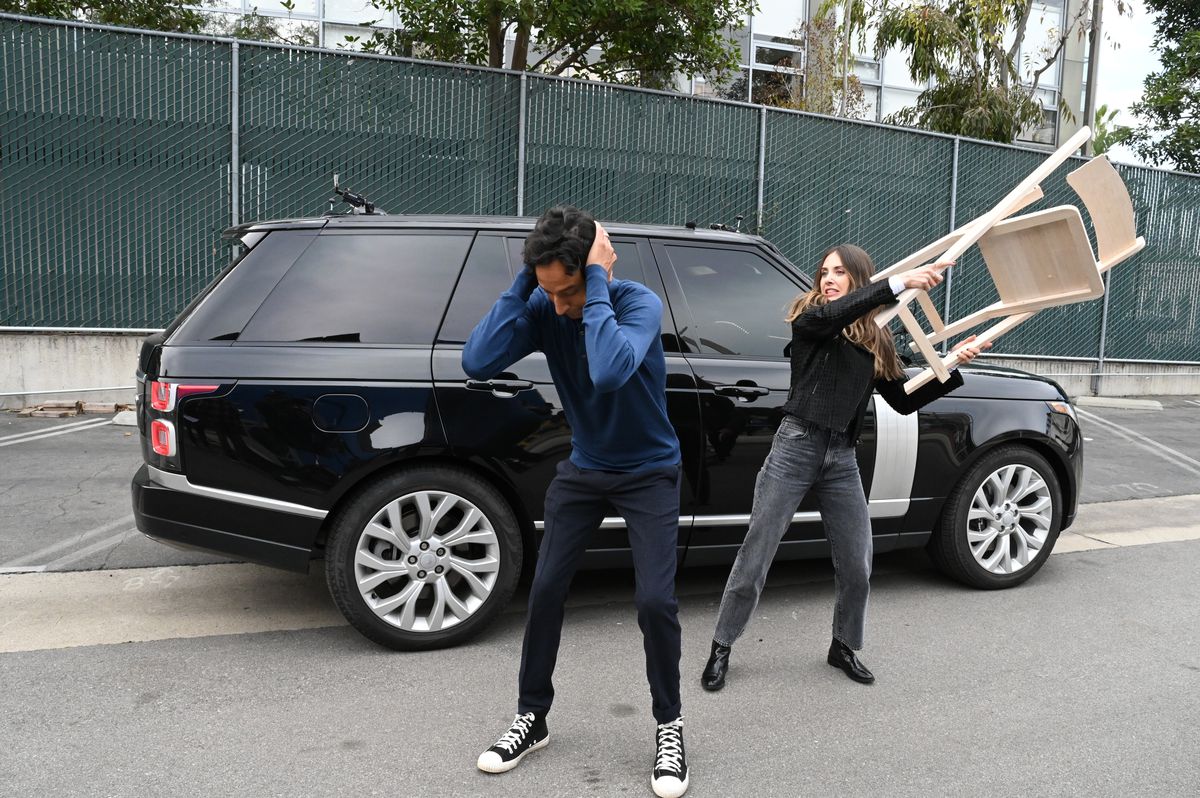 Danny Pudi håller för öronen och hukar lite medan Alison Brie kommer upp bakom honom med en stol på väg att slå honom.  Carpool Karaoke-bilen ligger bakom dem. 