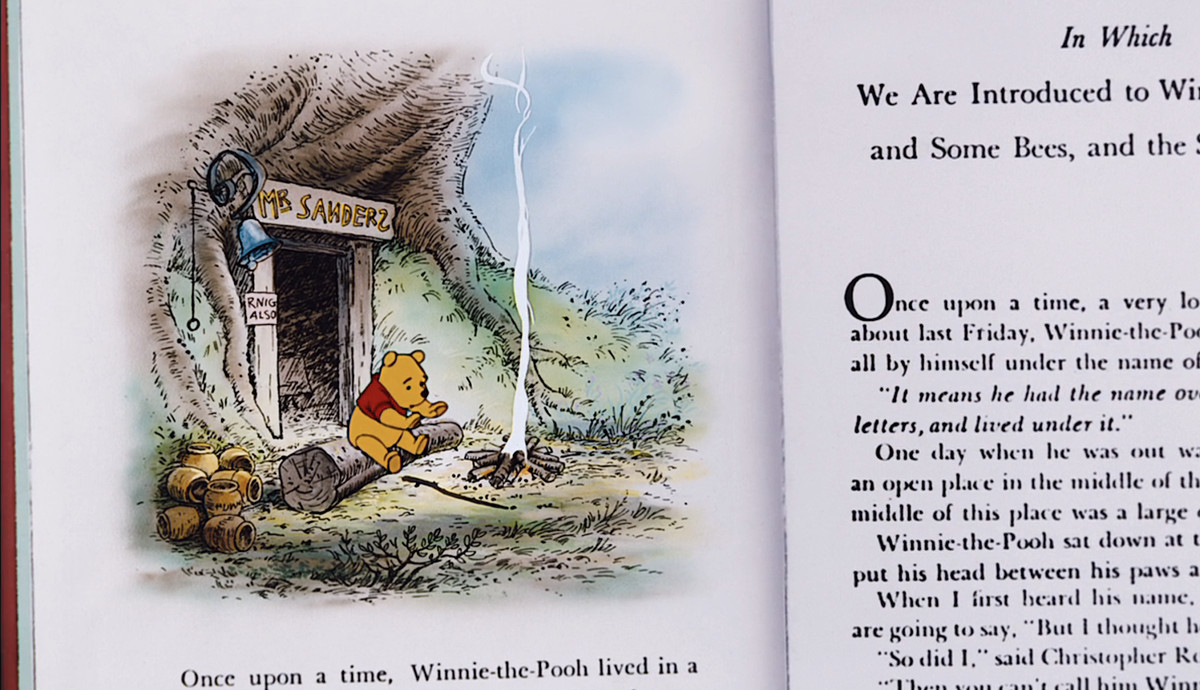 An image from The Many Adventures of Winnie the Pooh shows Pooh warming his paws over a small, smoky fire in front of his home built into the bottom of a tree, as an illustration within an animated book. Text below him: Once upon a time, Winnie-the-Pooh lived in a…”