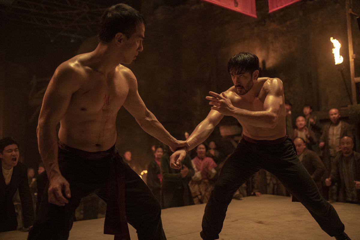 Andrew Koji and Joe Taslim, both shirtless and drenched in sweat, grasp onto each other in fighting stances as excited onlookers watch in Warrior.