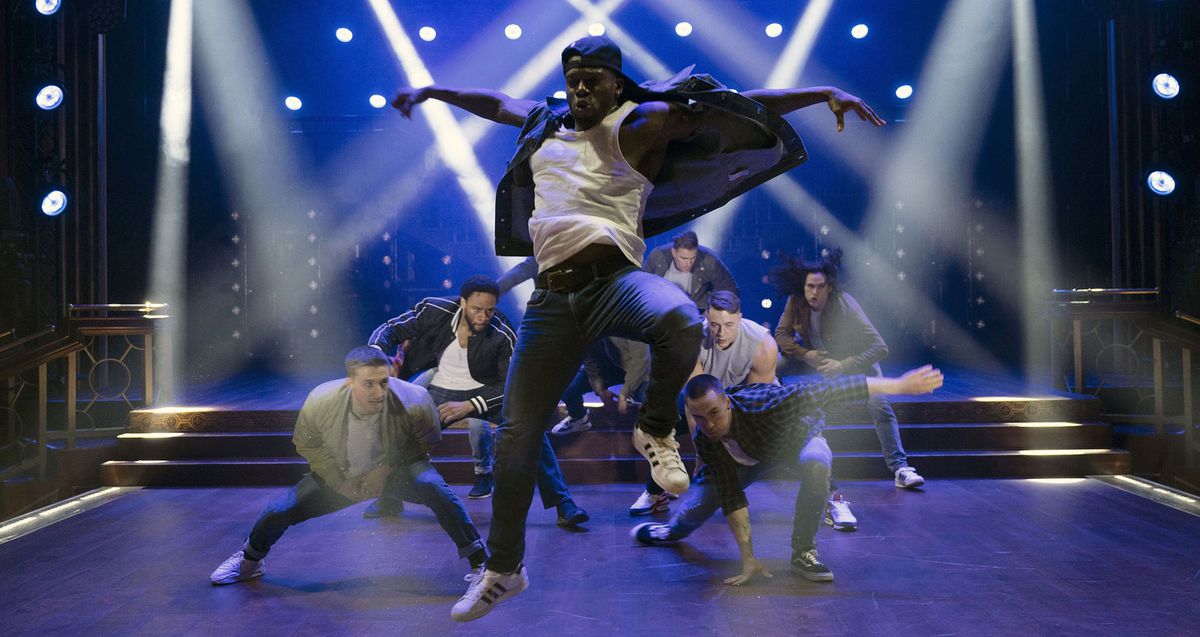 En svart manlig dansare i jeans och en bakåtvänd basebollkeps hoppar högt i luften när andra manliga dansare hukar bakom honom på en blåupplyst scen korsad med ljusa vita strålkastare i en scen från Magic Mike's Last Dance