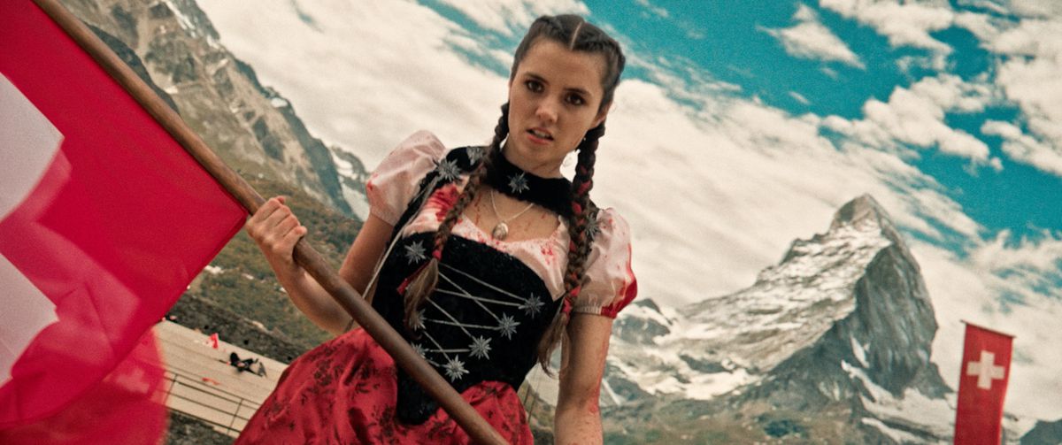 Alice Lucy som Heidi, klädd i en blodstänkt klänning, med en röd flagga med de schweiziska alperna i bakgrunden i Mad Heidi.