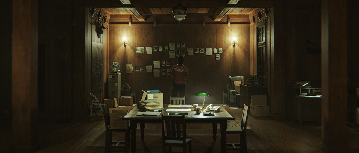 En bild av Saga som står i ett kontorsrum.  Det är svagt upplyst och det finns ett skrivbord fyllt med papper och en vägg med papper upptejpade i Alan Wake 2