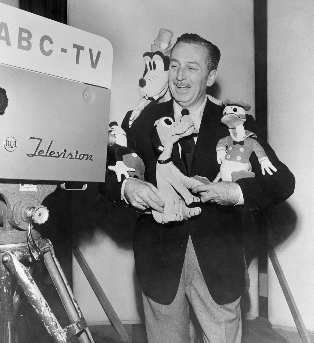 Walt Disney in an undated archive photo, standing in front of a camera labeled “ABC,” smiling and holding stuffed versions of Mickey Mouse, Donald Duck, and Pluto, with a stuffed Goofy looking over his shoulder