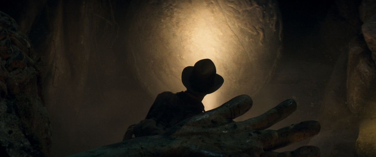 Silhuetten av Indiana Jones som utforskar en grav, handen på ett lik är i förgrunden, också skuggad.