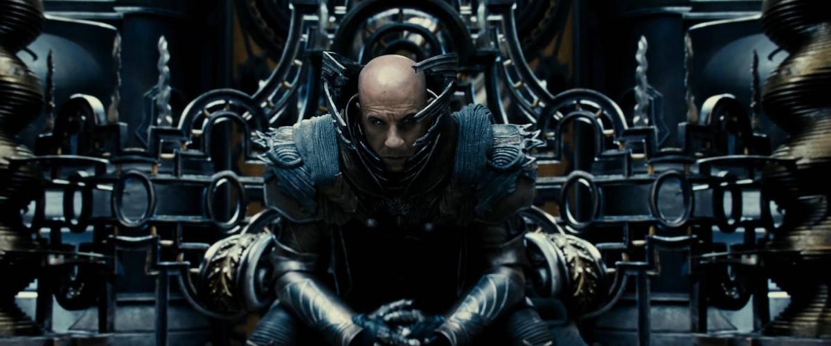 En skallig man (Vin Diesel) sitter på en tron, klädd i utarbetad metallisk rustning och omgiven av utarbetad teknologi utan tydligt syfte i The Chronicles of Riddick