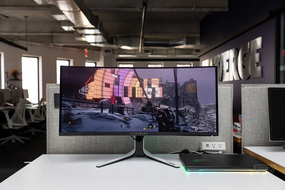 En bild av Alienware AW3423DW böjda spelmonitor på ett vitt skrivbord, ansluten till en bärbar speldator.