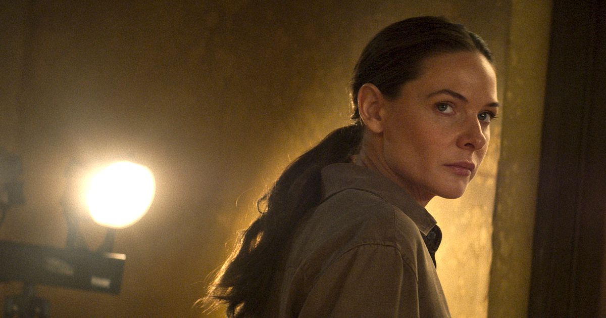 Ilsa Faust (Rebecca Ferguson), hår som är tillbakadraget i en hästsvans, står i ett obeskrivligt gulbrunt rum med ett starkt ljus bakom sig i Mission: Impossible – Dead Reckoning Part One