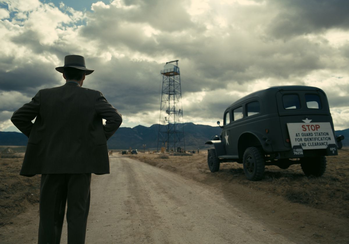 J. Robert Oppenheimer, i hatt, händerna på höfterna, står med ryggen mot kameran och betraktar tornet vid testplatsen för atombomben framför en enorm, molnig ökenhimmel i filmen Oppenheimer