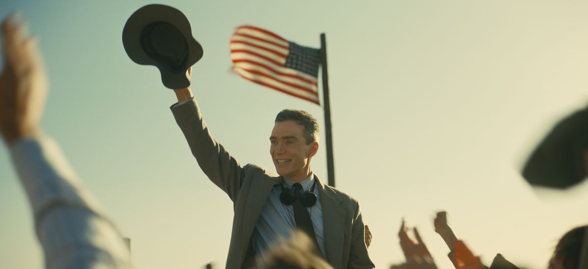 J. Robert Oppenheimer (Cillian Murphy) flinar och viftar med hatten mot en jublande utomhuspublik medan han står under en amerikansk flagga i Christopher Nolans Oppenheimer
