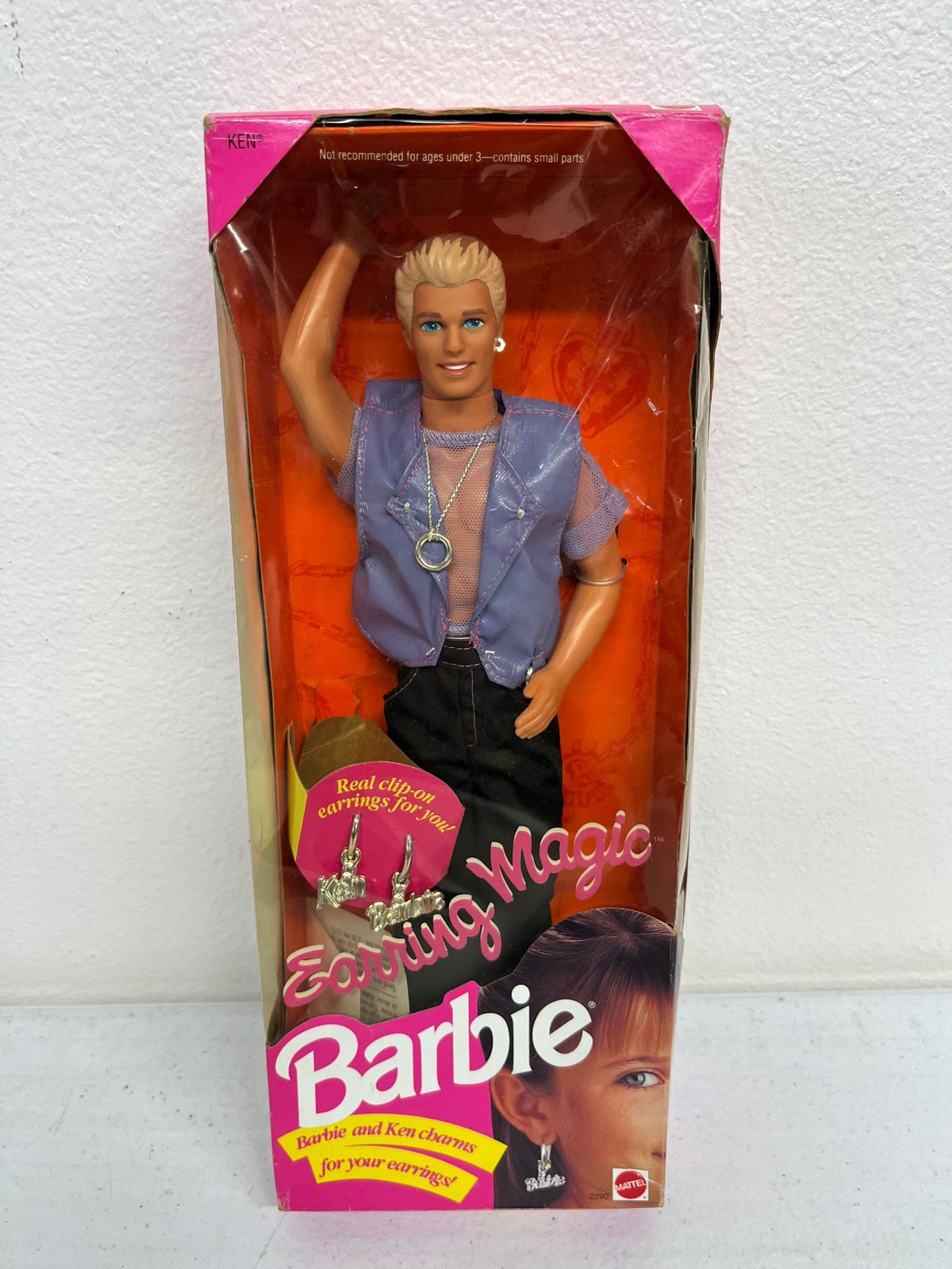 En produktbild av Earring Magic Ken i kartongen: En Ken-docka med brunt och blont streck hår, en lila meshskjorta och lila läderväst och en mycket synlig silverring på en kedja runt halsen