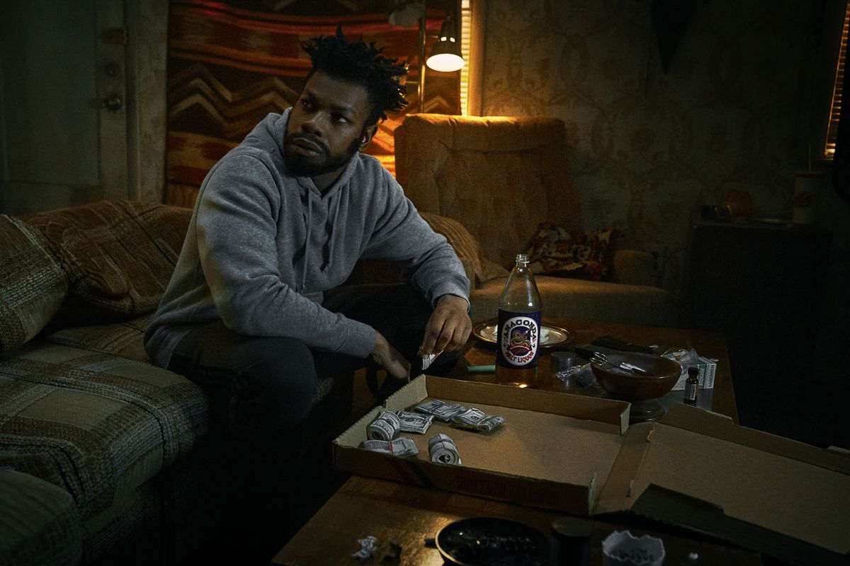 John Boyega som Fontaine sitter på en soffa framför en pizzakartong fylld med rullar med dollarsedlar i They Cloned Tyrone.