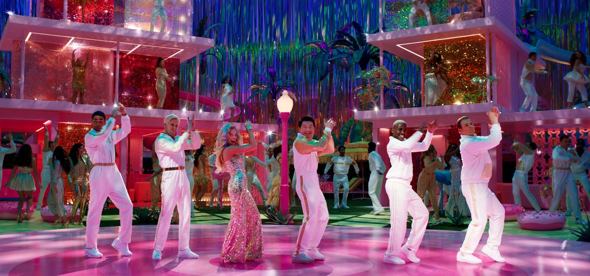 Barbie (Margot Robbie), i en glittrig rosa klänning, dansar linedance framför ett par vägglösa rosa Barbie Dreamhouses i naturlig storlek, flankerade av fem Kens i helvitt, spelade av Kingsley Ben-Adir, Ryan Gosling , Simu Liu, Ncuti Gatwa och Scott Evans, i filmen Barbie från 2023