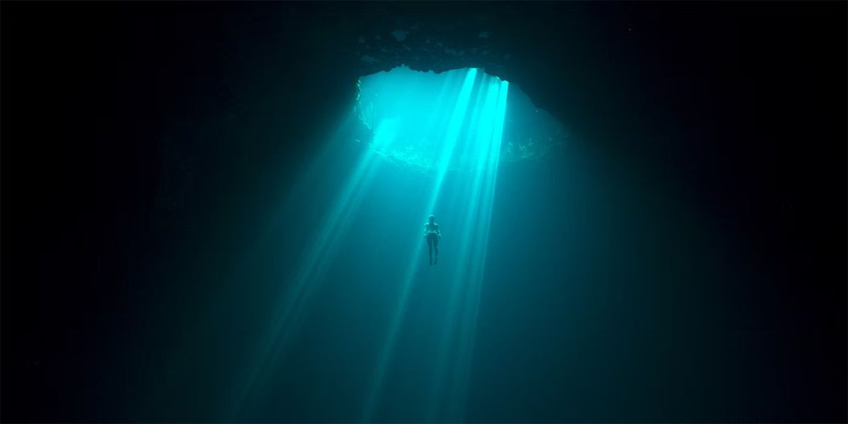 En ensam figur som stiger ner i ett massivt hål i havet omgiven av mörker, upplyst i en ljusstråle i The Deepest Breath.