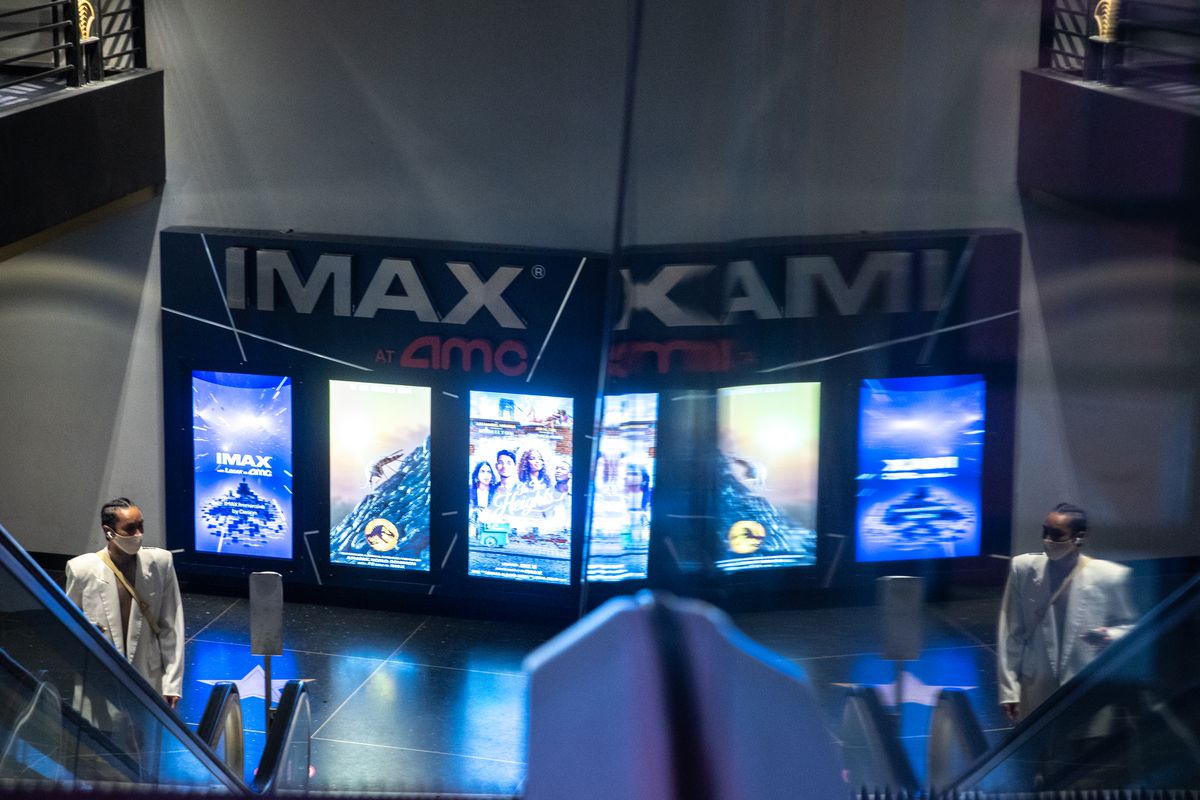 A masked woman rides an escalator up in an AMC theater, with a giant IMAX sign and a series of backlit movie posters behind her, and reflected in a mirrored wall next to her