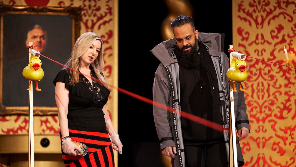 Victoria Coren Mitchell och Guz Khan tittar förvirrade på en rad gummiänder på snören i Taskmaster-studion.