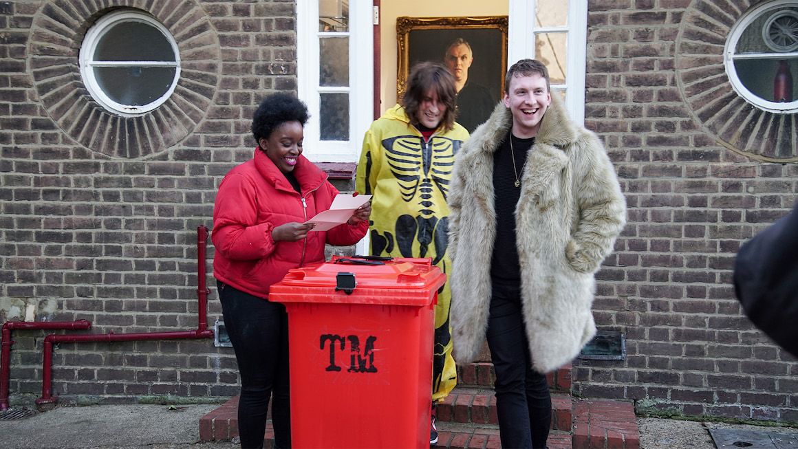 Lolly Adefope, Noel Fielding och Jo Lycett ler tillsammans medan de läser en uppgift ovanför en röd papperskorg utanför Taskmaster-huset.  Adefope bär en knallröd rock, Fielding bär en skelettdräkt och Lycett bär en stor päls.