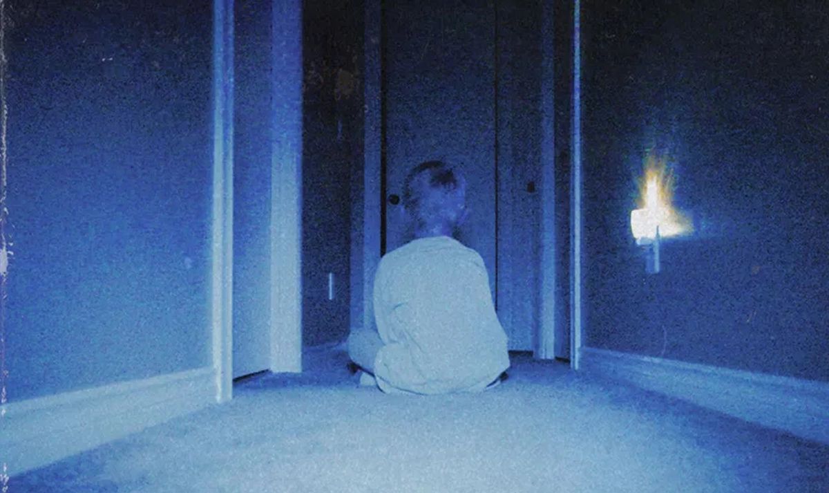 A young boy sits in a dim, blue hallway with his back to the camera, facing a series of open doorways, in a typically grainy, fuzzy shot from the horror movie Skinamarink