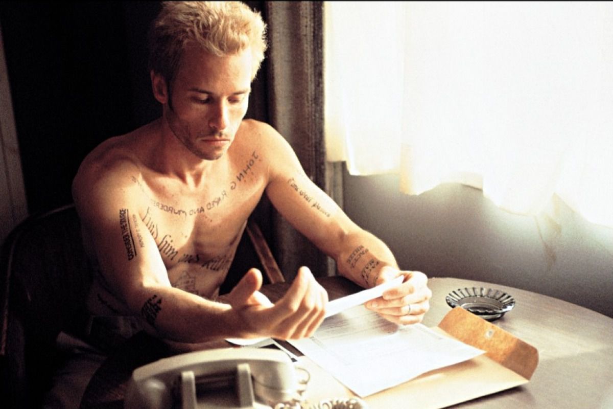 Leonard Shelby (Guy Pearce) sitter bar överkropp framför ett gardinfönster på ett hotellrum, täckt av påminnelsetatueringar och med lite pappersarbete framför sig i Christopher Nolans Memento.