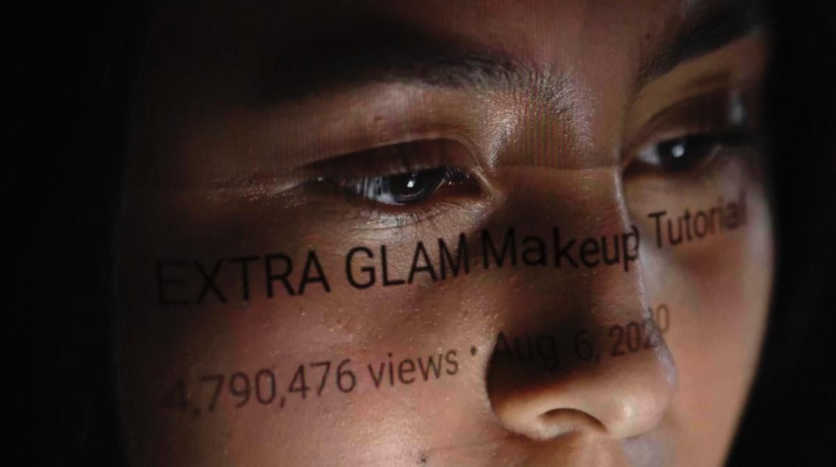 En ung persons ansikte som orden ”EXTRA GLAM MAKEUP Tutorial;  4 790 476 visningar;  2020” spelar över deras ansikte som en reflektion av skärmen.