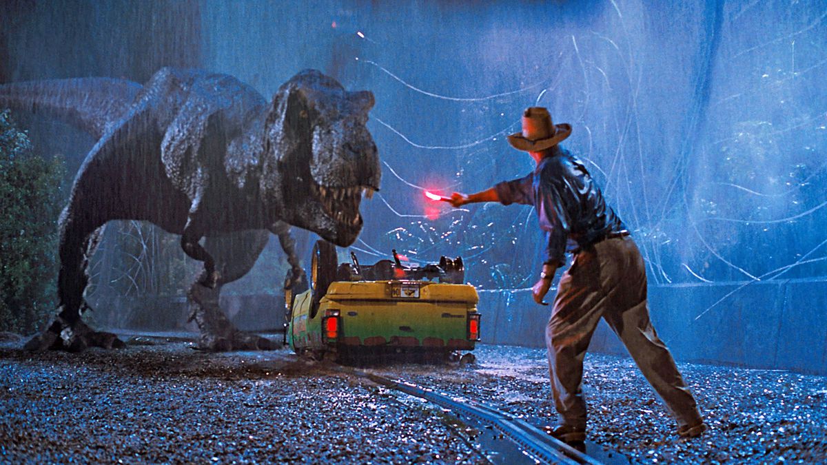 En man i brun hatt och blå skjorta (Sam Neill) viftar med en tänd nödsignal för att distrahera en Tyrannosaurus Rex som står bredvid en vältad grön och gul lastbil bredvid en trasig vajerport.