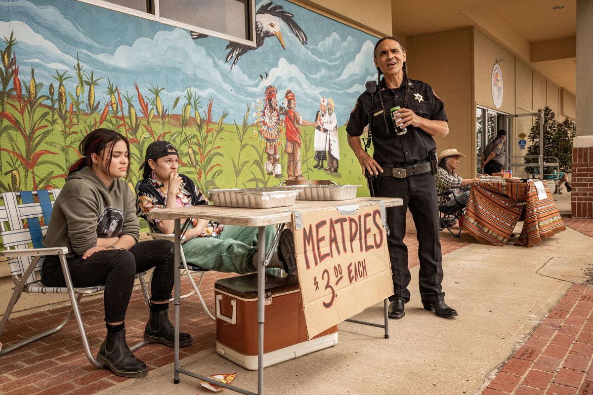 Devery Jacobs, Paulina Alexis och Zahn McClarnon samlas runt ett bord som annonserar Meatpies för $3 st, framför en väggmålning, i Reservation Dogs.