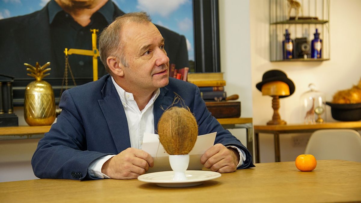 Bob Mortimer ser förbryllad ut när han sitter framför en kokosnöt i en äggkopp.  Han läser från ett vitt papper, och en klementin är bredvid honom, i Taskmaster Series 5.