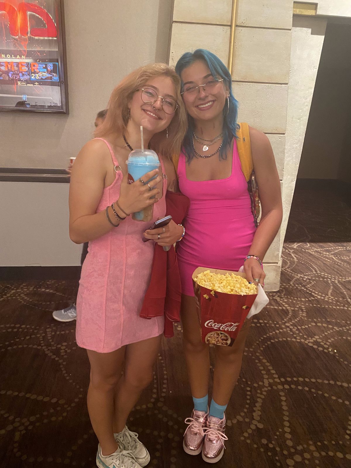 Två personer i rosa klänningar står i en biograf medan de håller i popcorn och en drink.