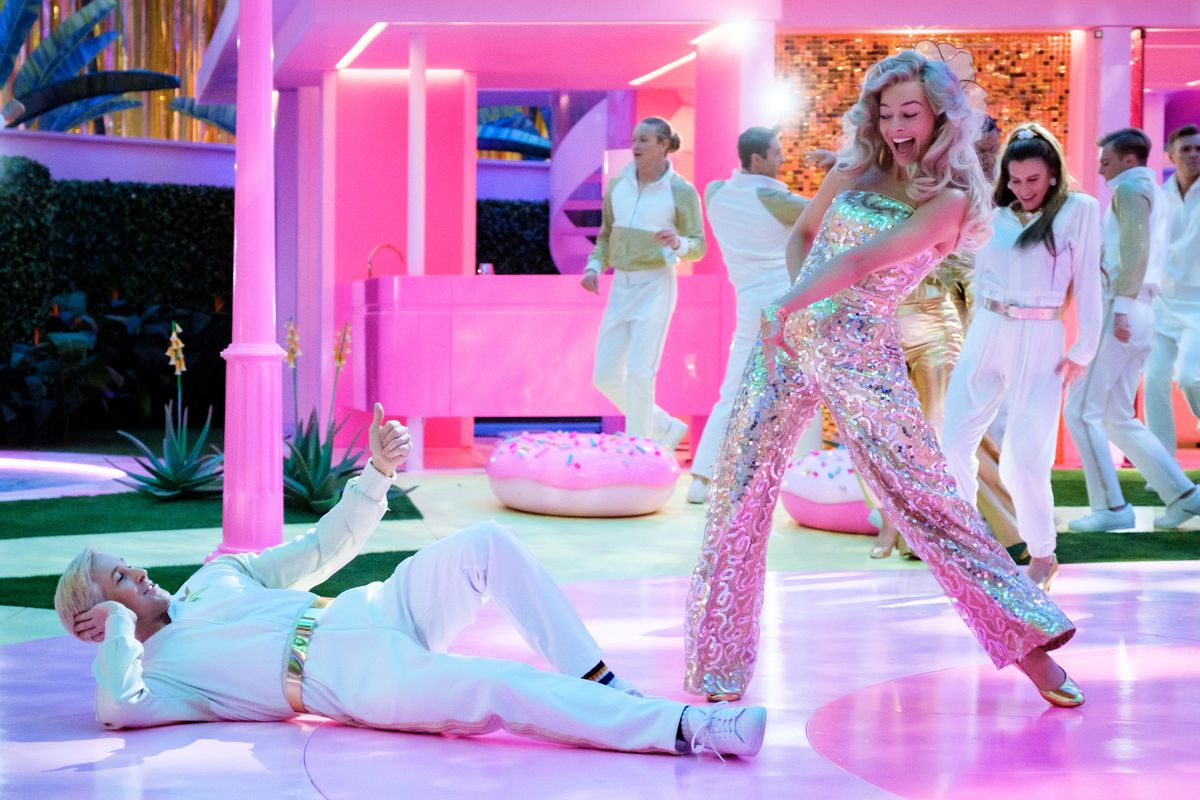 Barbie (Margot Robbie), i en silverfärgad jumpsuit, flinar mot Ken (Ryan Gosling), i en helvit outfit, liggande på ett glänsande rosa dansgolv med andra dansande Barbies och Kens i bakgrunden i Greta Gerwigs live-action 2023 filmen Barbie