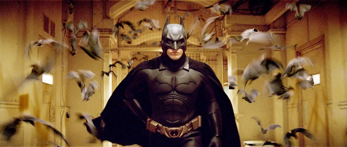 Batman (Christian Bale) går genom en gulaktig korridor, omgiven av en flock suddiga, snabbrörliga fladdermöss i Christopher Nolans Batman Begins