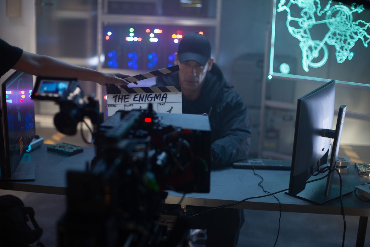 Ett foto bakom kulisserna av John Cena som filmar sitt framträdande som The Enigma.  John sitter vid ett skrivbord, flankerad av bildskärmar och datorer, med ett serverställ bakom sig.