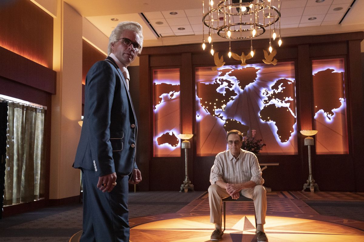 John Goodman sitter i en stol i The Righteous Gemstones tronrum, ovanpå en kompass och framför en upplyst global karta.  Walton Goggins går framför honom i en blå kostym.
