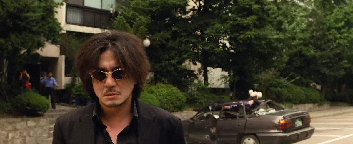 Oh Dae-su (Choi Min-sik), en lurvig man med solglasögon och svart skjorta, står på gatan och ser uttryckslös ut, framför en bil som har krossats av en fallande människokropp i Park Chan-wooks Oldboy