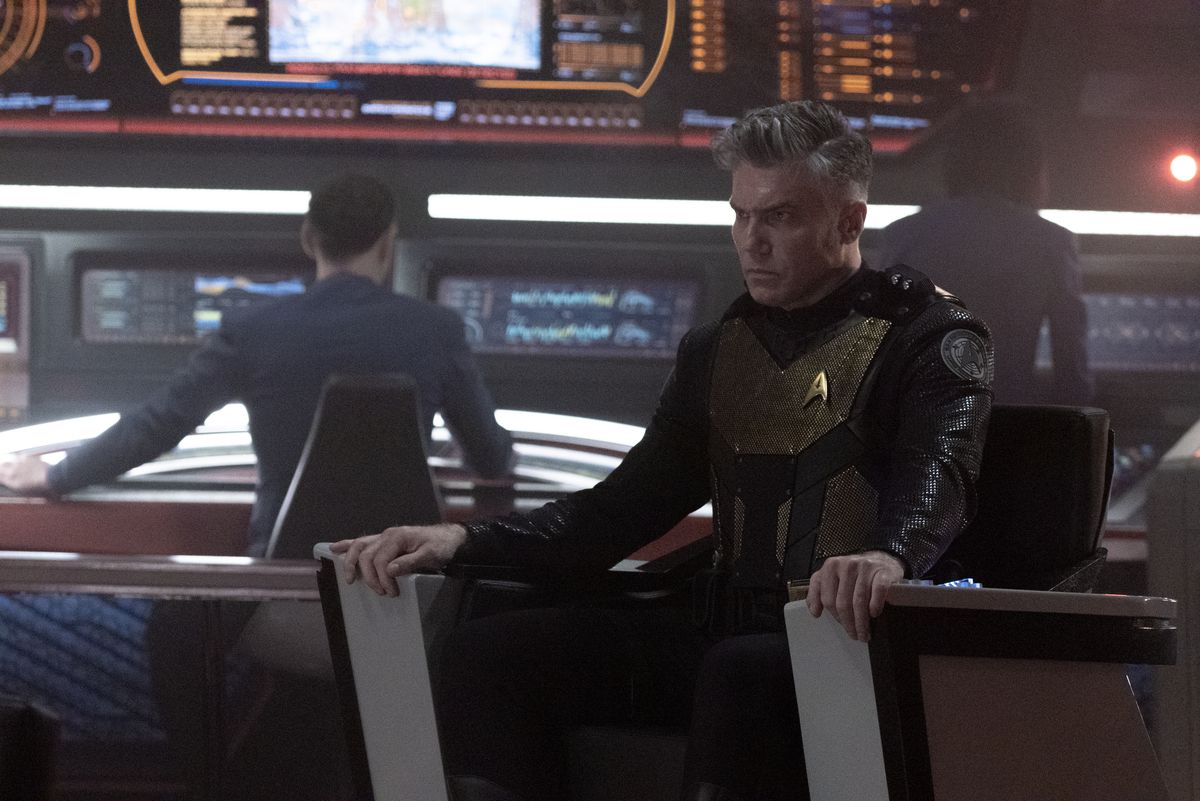 Anson Mount som kapten Pike sitter strängt i sin kaptensstol, iklädd taktisk rustning, med Ethan Peck som Spock synlig vid sin konsol i bakgrunden, i Star Trek: Strange New Worlds.