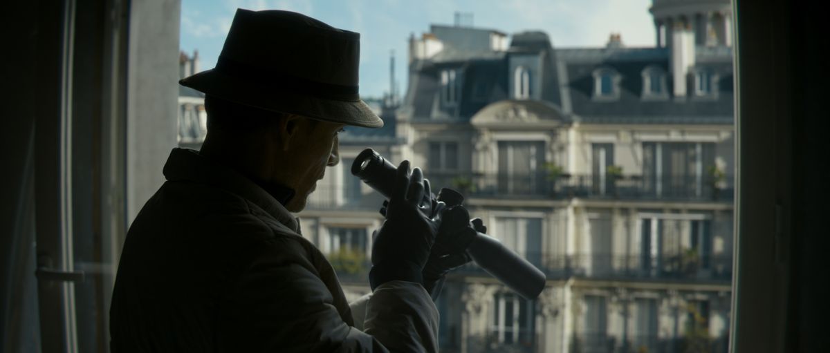 Michael Fassbender looks out a window with a mini telescope, like the kind you’d put on a rifle, in The Killer.