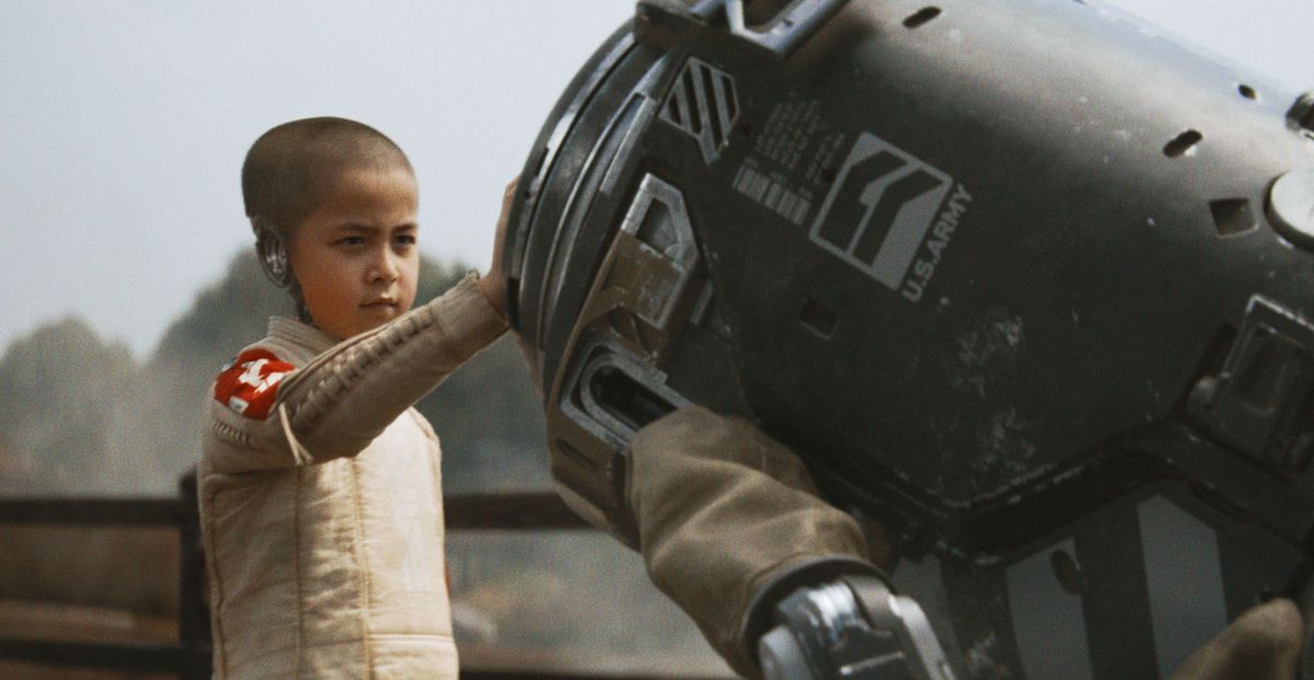 Ett ungt androidbarn med ett rakat huvud och delvis saknad skalle lägger sin hand på ett maskineri i The Creator