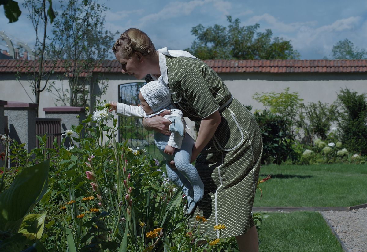 A woman leans over some flowers to let the baby she’s holding touch them in The Zone of Interest.