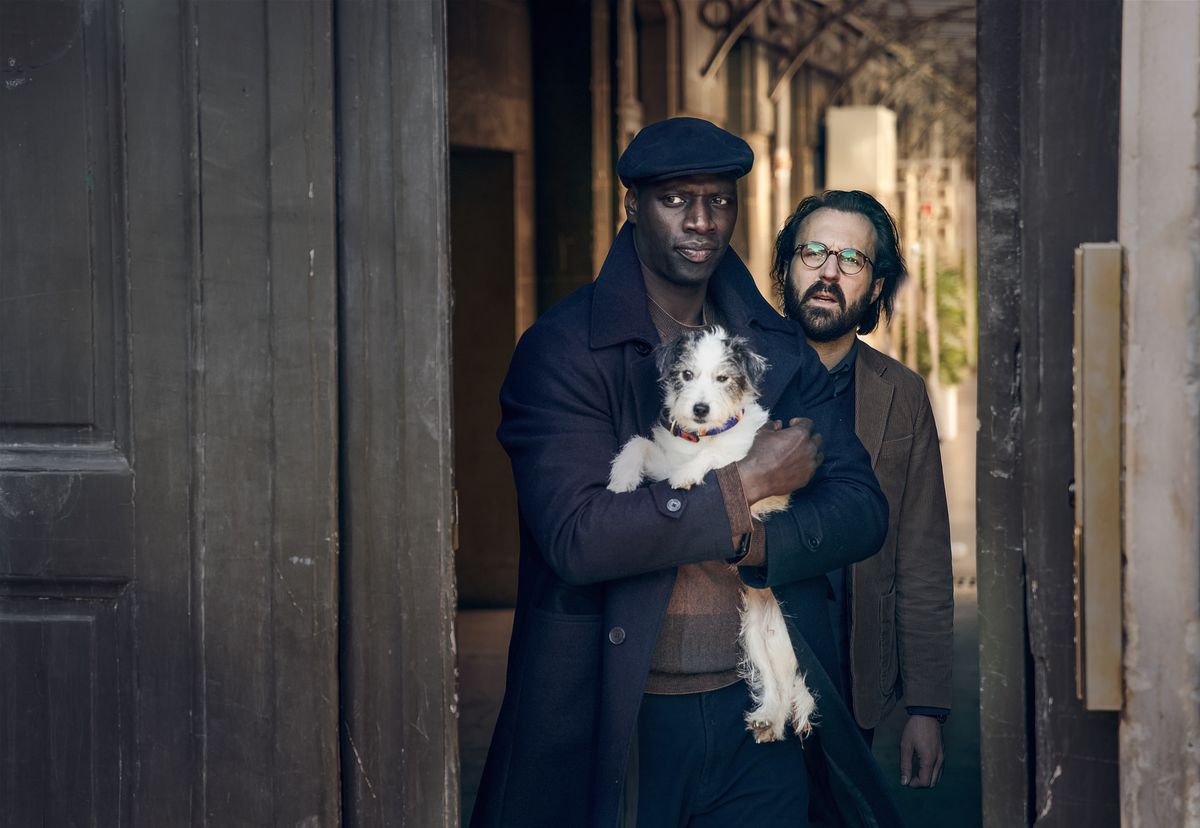 Omar Sy as Lupin holds an adorable dog as he walks through a doorway with another man.