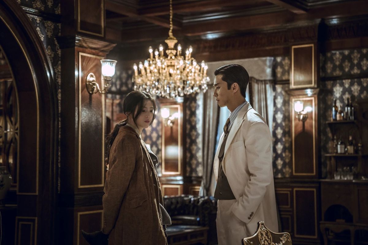 Park Seo-joon och Han So-hee bär fina kläder och står bredvid varandra i ett utsmyckat rum med en stor ljuskrona i Gyeongsang Creature.