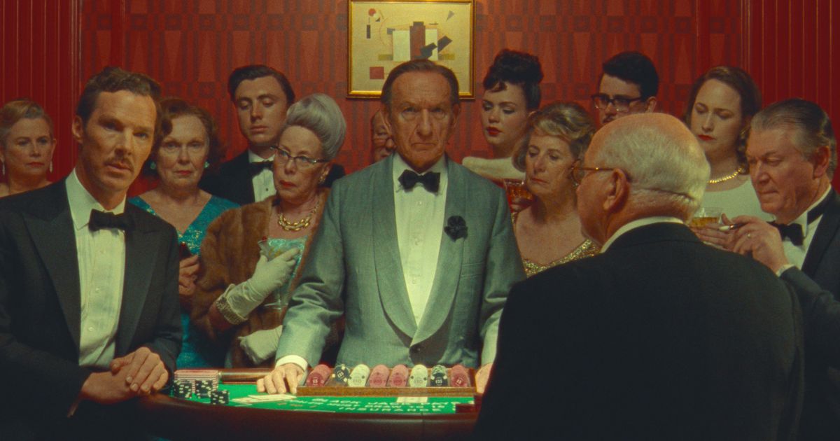 Benedict Cumberbatch (i smoking som Henry Sugar) och Sir Ben Kingsley (som croupier) tittar in i kameran när de står vid ett bord på ett kasino, omgivna av en nyfiken skara välklädda människor, i Wes Andersons Netflix-film The Underbar berättelse om Henry Sugar
