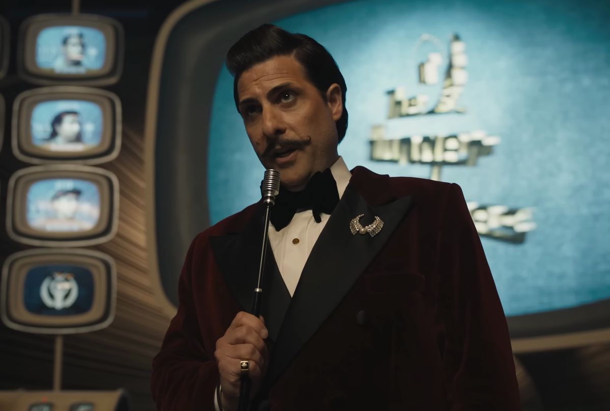 Jason Schwartzman med styrets mustasch spelar Lucky Flickerman och håller en mikrofon framför Hunger Games TV i The Ballad of Songbirds and Snakes