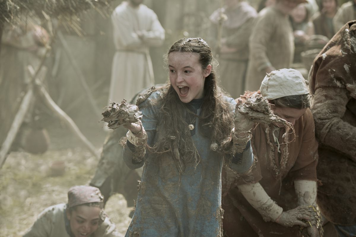 a young teenage girl with pale skin and long brown hair looks excited as she grabs a fistful of mud in each hand