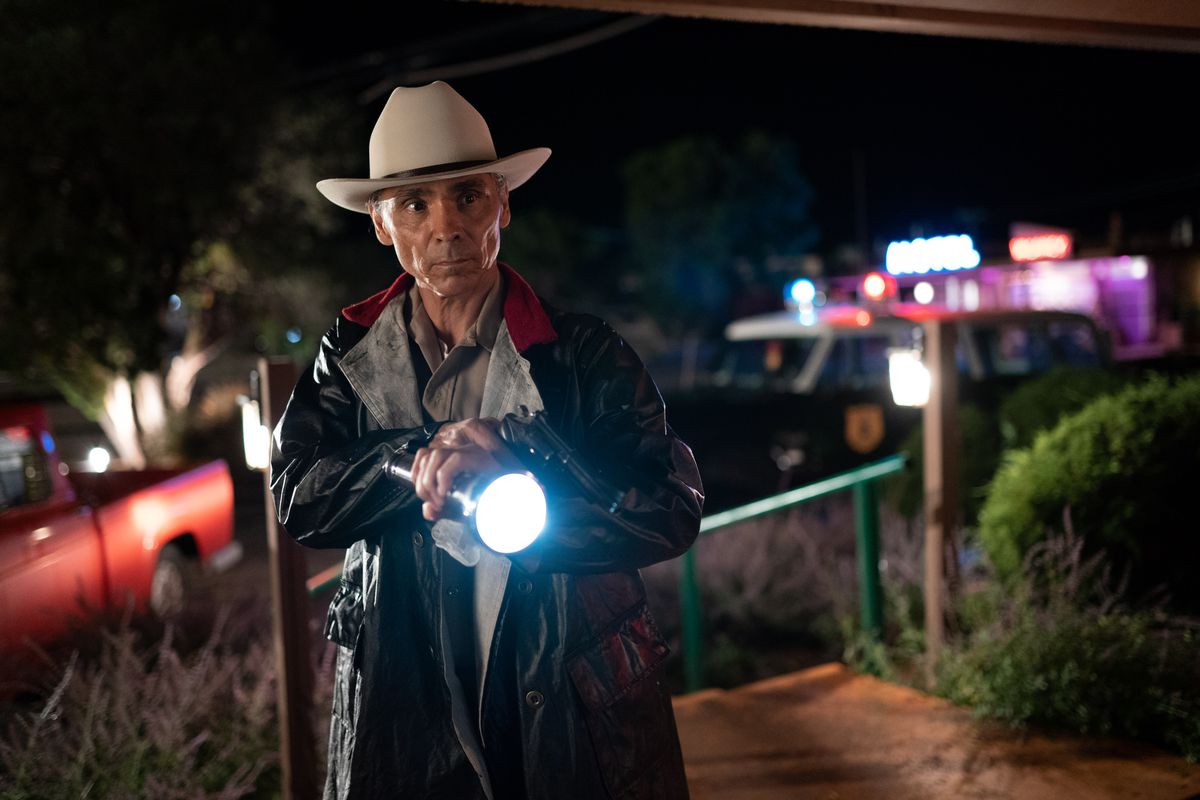 Zahn McClarnon som officer Joe Leaphorn höjer en ficklampa och en pistol på en ökennatt med neonljus bakom sig i AMC:s Dark Winds.