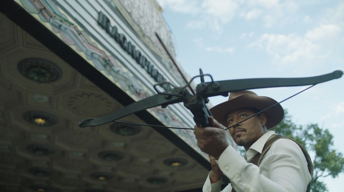 Terrence Howard håller ett armborst framför ett filmtält medan han bär en cowboyhatt i Showdown at the Grand.