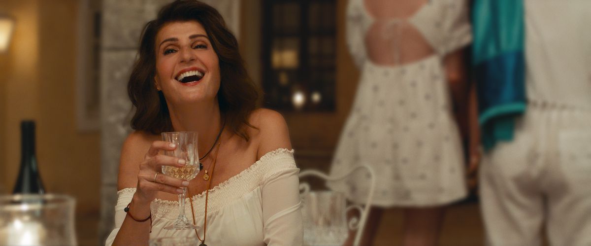 Toula skrattar och höjer ett glas i en vit sommarklänning under ett bröllop i My Big Fat Greek Wedding 3