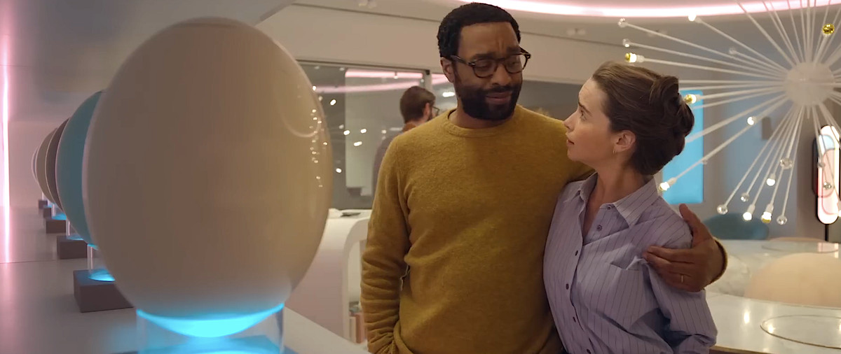 Alvy (Chiwetel Ejiofor) ler och kramar sin fru Rachel (Emilia Clarke) när de står i ett ljust pastellfärgat utrymme fullt av enorma, glänsande plastägg som visas till försäljning i The Pod Generation